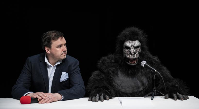 Man in a suit sitting beside a fake gorilla.
